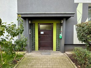 Ein Gebäudeeingang mit einer dunkelbraunen Tür, grünen vertikalen Akzenten und einem kleinen Fenster mit einem Metallgitter.