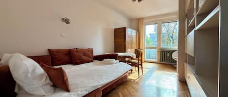 A hotel room with a bed, a wooden floor, a dining table, and a view of the outside.