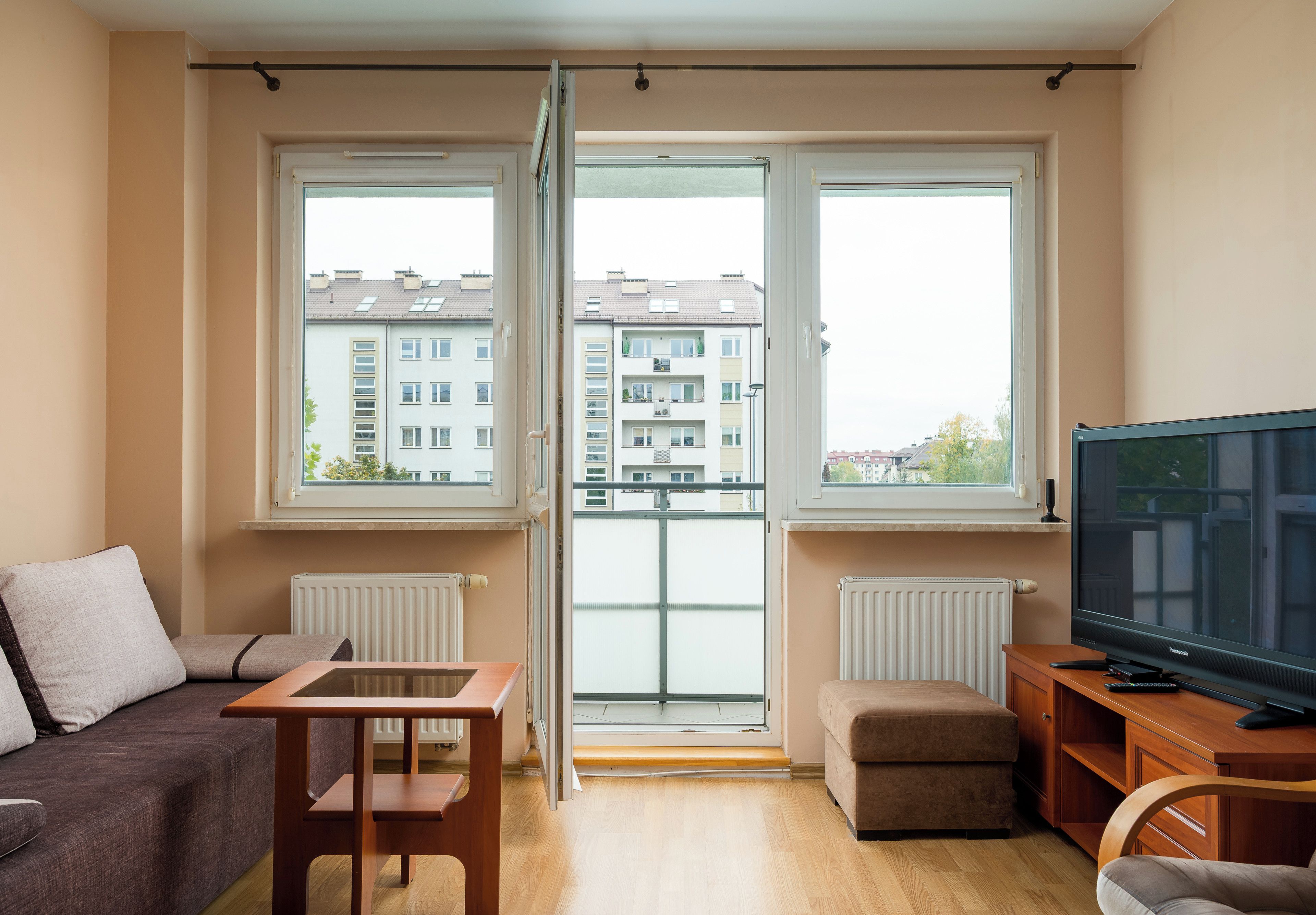 En moderne stue med sofa, et lille bord, fjernsyn og en balkon med udsigt over boligbygninger.