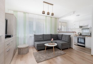 Een moderne woonkamer met een grijze hoekbank, een kleine ronde tafel met een potplant, een keuken met een fornuis en oven, en lichte gordijnen voor de ramen.