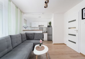 A modern living room with a grey sofa, a small white table with a plant, and a kitchen in the background. - Osiedle na Stoku Comfort Studio (Kraków)