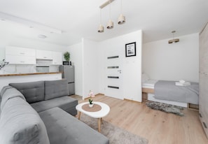 A modern living space with a grey sofa, a small round table with a plant, a bed with a grey quilt, and a kitchen area with white cabinets and a microwave. - Osiedle na Stoku Comfort Studio (Kraków)