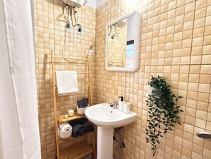 A bathroom with a tiled wall, a white sink, a wooden shelf with towels and toiletries, a mirror, and a hanging plant. - New Listing (Puerto del Rosario)