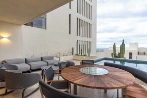 A modern outdoor patio with a round glass table, grey sofas, and a pool. - Modern Ocean View 2BR Condo Resort-Style Pool, Gym & Prime Cabo Location (Cabo San Lucas)