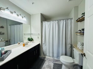 A bathroom with a shower curtain, a toilet, a sink with a mirror, and a shelf with toiletries. - Spacious 4-bedroom townhome with AC in peaceful Kissimmee FL Gated Community, (Kissimmee)