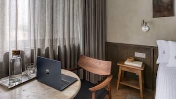 A hotel room with a laptop on a round table, a wooden chair, a small wooden side table with a book and a bowl, and a bed with white bedding.