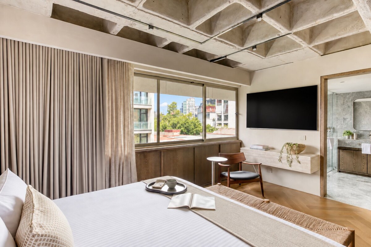 Una habitación de hotel moderna con una cama grande, una mesita, una silla, un televisor y un baño con espejo y lavamanos.