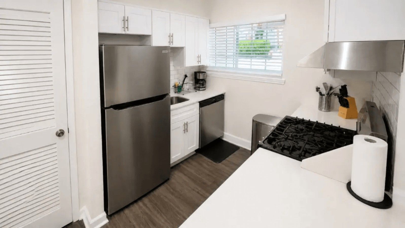 A modern kitchen with white cabinets, a stainless steel refrigerator, a gas stove, and a window with blinds.