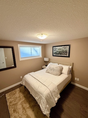 A bedroom with a bed, a nightstand, a lamp, a window with blinds, a framed picture, and a ceiling light. - Modern & Cozy 2BR | Near Refinery, Airport & Central Regina (Regina)