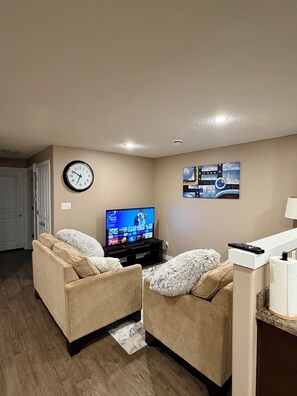 Uma sala de estar com dois sofás bege, uma televisão, um relógio e um quadro na parede.