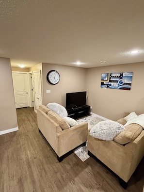 A living room with two sofas, a flat-screen TV, a clock, and a painting on the wall. - Modern & Cozy 2BR | Near Refinery, Airport & Central Regina (Regina)