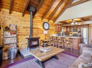 A cozy cabin interior with a wood-burning stove, a kitchen with modern appliances, and a bar area. - Cozy Big Bear Cabin • Sleeps 10 • Pet Friendly! (Big Bear)