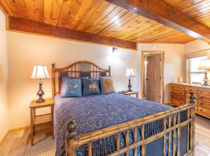 A bedroom with a wooden ceiling, a bed with a blue patterned bedspread, two bedside lamps, and a wooden dresser. - Cozy Big Bear Cabin • Sleeps 10 • Pet Friendly! (Big Bear)