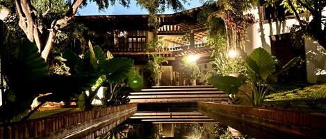 A multi-level wooden structure with a central staircase, surrounded by lush greenery and a reflective water feature.