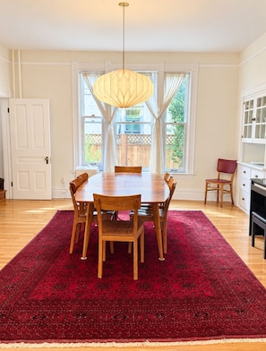 A dining area with a wooden table and chairs, a red rug, a hanging light fixture, and a view of the outdoors through large windows. - Historic 4BR and 3 Bath, University District Home, Sleeps 8, Near UM & Downtown (Missoula)