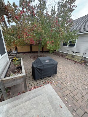 A patio with a grill cover, a wooden planter box with plants, and a tree with autumn leaves. - Historic 4BR and 3 Bath, University District Home, Sleeps 8, Near UM & Downtown (Missoula)