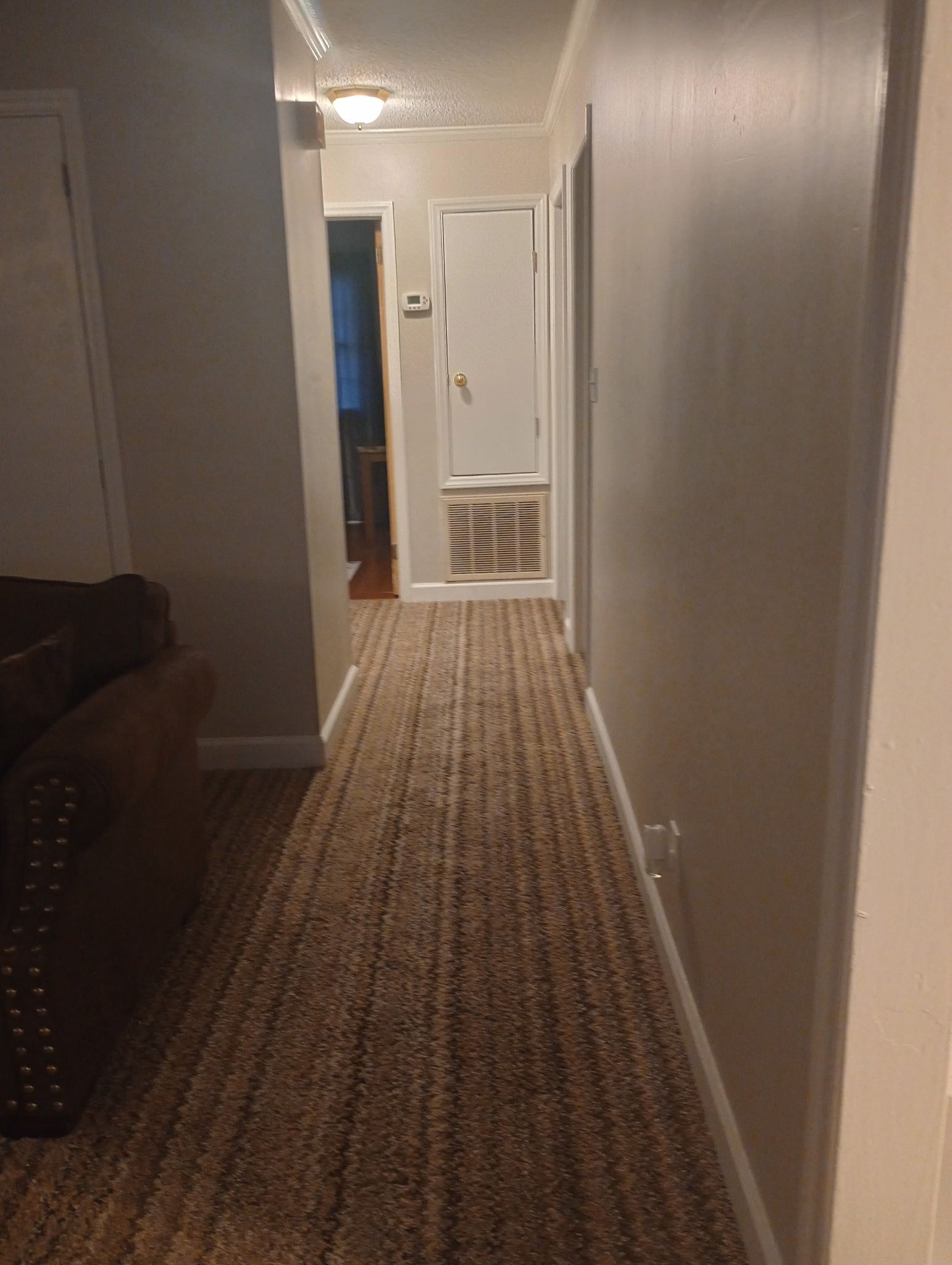 Ein Teppich ausgelegter Flur mit einer Tür am Ende, einem Thermostat an der Wand und einem Einblick in einen Raum mit einem braunen Sofa.