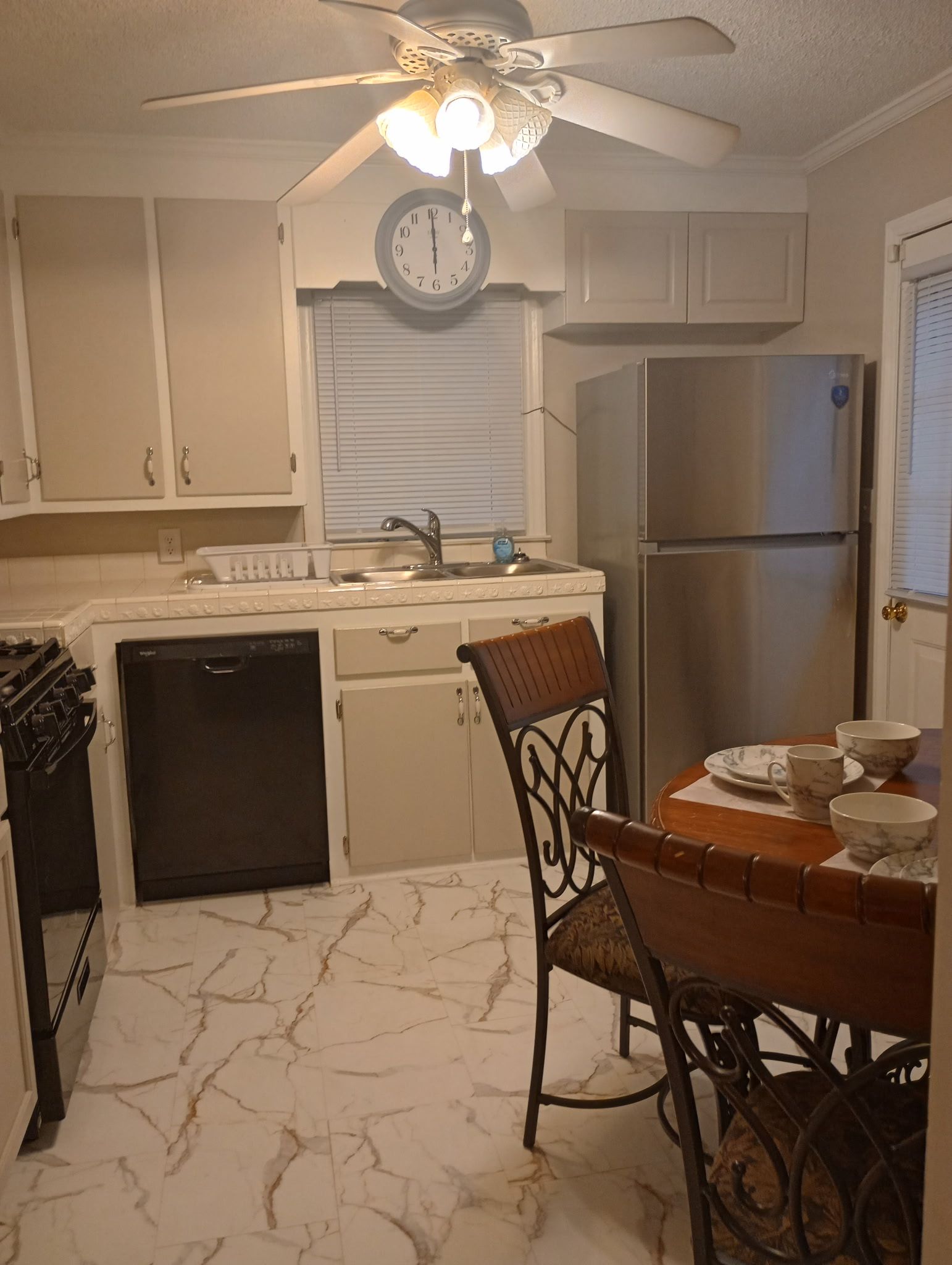 A kitchen with a clock, a refrigerator, a stove, a dishwasher, and a dining table with chairs.
