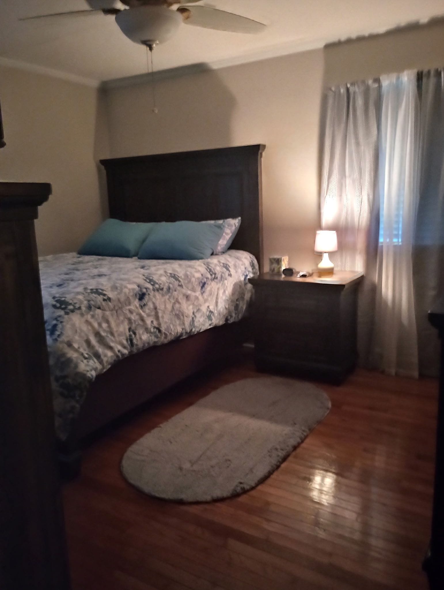 Un dormitorio con cama, mesita de noche con lámpara, ventilador de techo y ventana con cortinas.