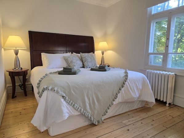 A neatly made bed with a headboard, two bedside lamps, a radiator, and a window with a view of trees.