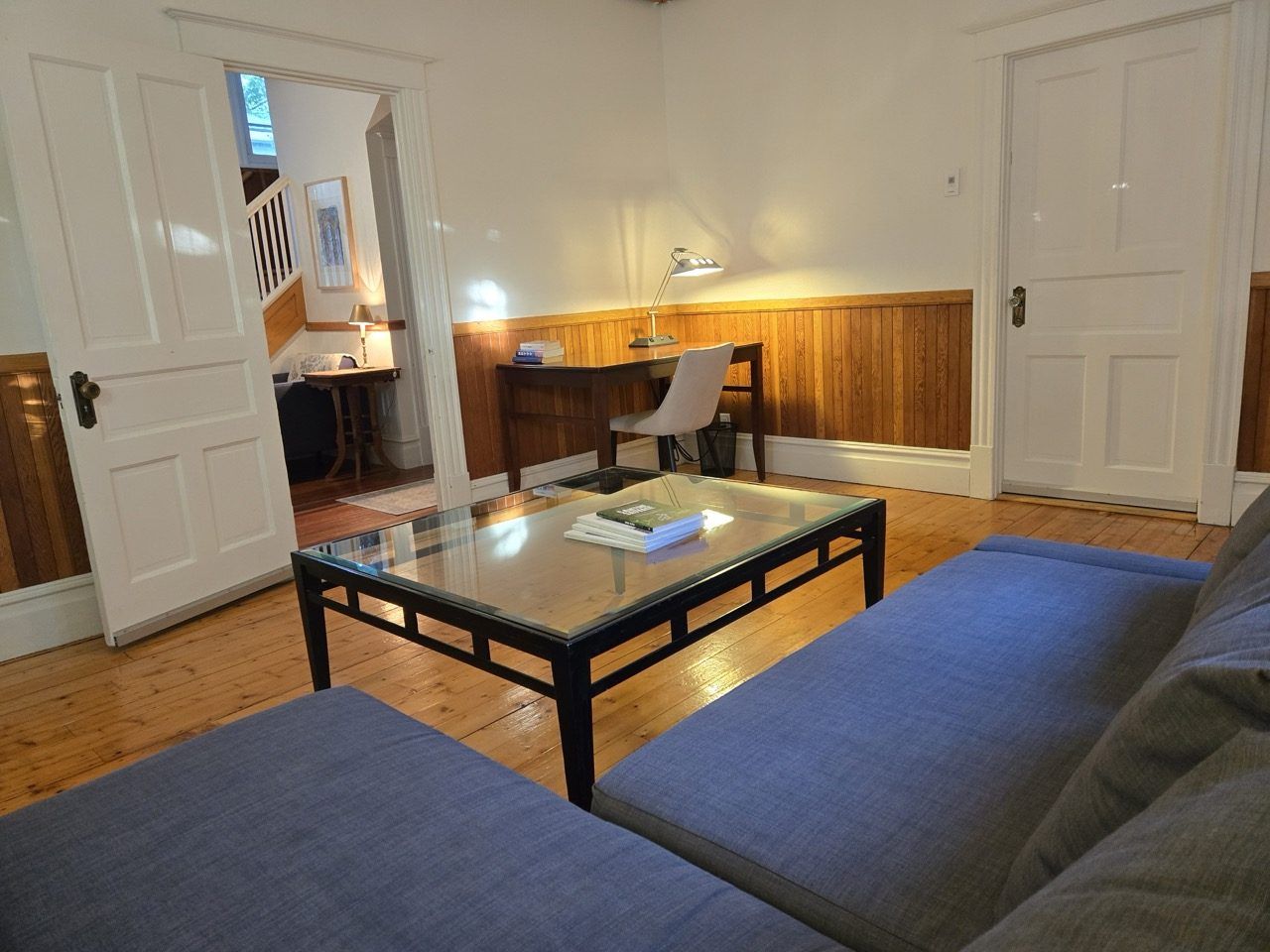 A room with a glass-top coffee table, a blue sofa, a wooden desk, and a chair.