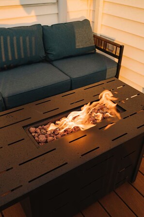 Une table en métal dotée d’un foyer intégré, un canapé de couleur sombre avec des coussins et une chaise en bois.