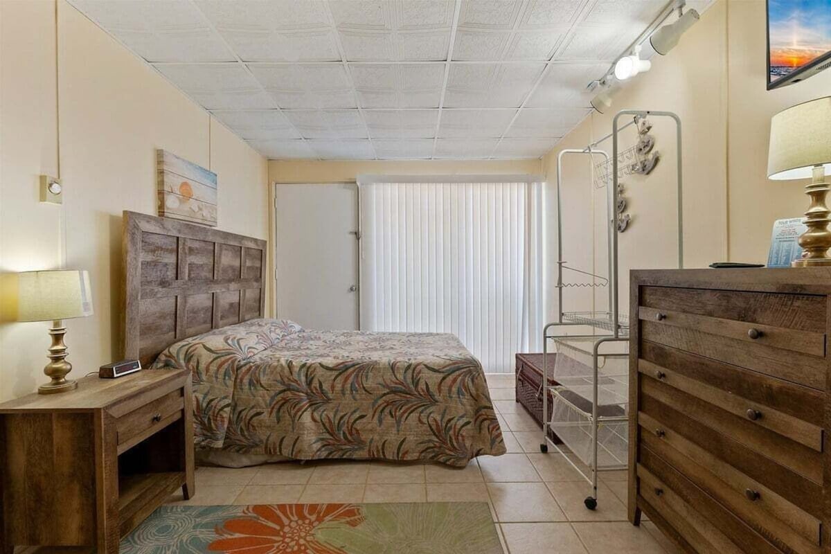 Een slaapkamer met een bed, een dressoir, een nachtkastje, een lamp, een raam met jaloezieën en een deur.