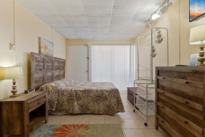 Een slaapkamer met een bed, een dressoir, een nachtkastje, een lamp, een raam met jaloezieën en een deur.