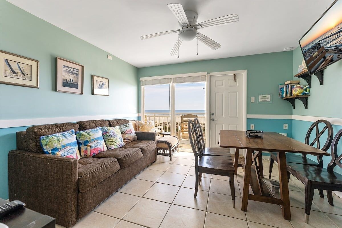 A living room with a sofa, a dining table, chairs, a ceiling fan, and a view through a glass door.