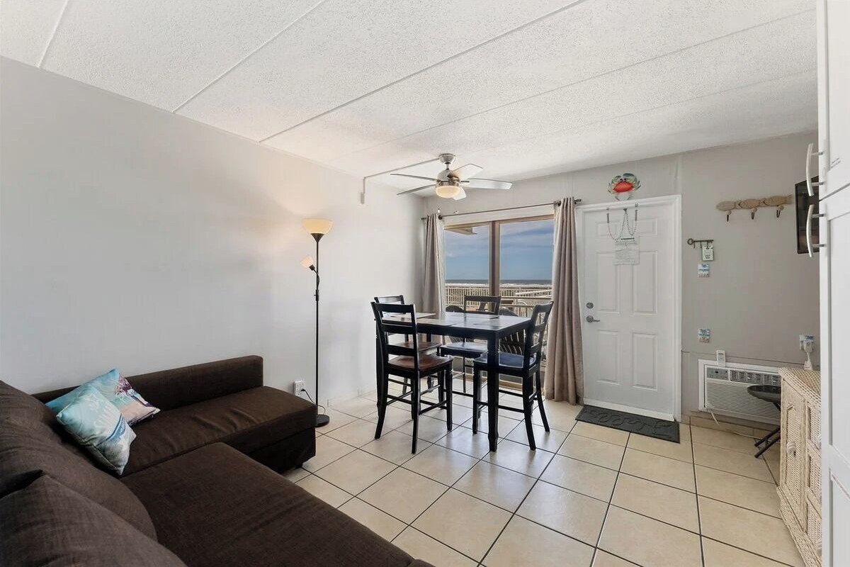 A living room with a sofa, a dining table with chairs, a ceiling fan, and a view through a door.