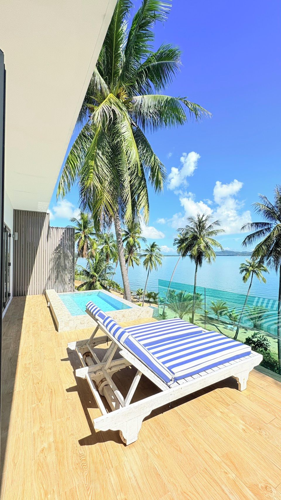 A poolside area with a lounge chair and a pool, surrounded by palm trees and a view of the ocean.