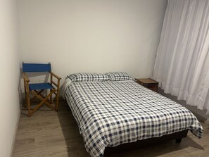 A single bed with a checkered bedspread, a wooden folding chair, a nightstand, and a window with curtains. - New Listing (Bogotá)
