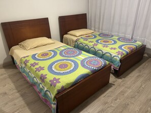 Dos camas de madera con ropa de cama colorida, suelo de madera y una cortina blanca.