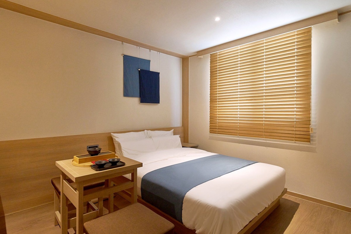 A hotel room with a bed, bedside table, and a window with blinds.