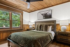 A bedroom with a wooden ceiling, a large bed with a quilted green bedspread, a headboard with multiple pillows, a window with a view of trees, and a bull-themed artwork on the wall.