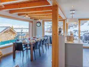 A dining area with a long table set for a meal, wooden beams on the ceiling, and large windows offering a view of a snowy landscape. - Nice apartment in villa for 8 guests with WIFI, TV and terrace (Basse-nendaz)