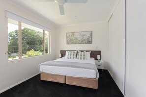 Ein Schlafzimmer mit einem großen Bett, einem Nachttisch, einem Deckenventilator und einem Fenster mit Blick auf Grünflächen.