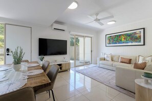 A modern living room with a flat-screen TV, a ceiling fan, a sofa, and a dining area with a table set for four. - Escape on St Crispins Pet Friendly Retreat (Port Douglas)