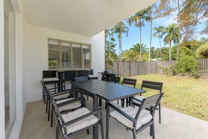 Eine Terrasse im Freien mit Esstisch, Stühlen, einem Grill und Blick auf einen Garten mit Palmen.