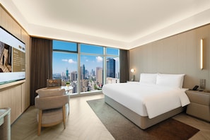 A modern hotel room with a large bed, a desk, a chair, and a view of a cityscape through floor-to-ceiling windows.