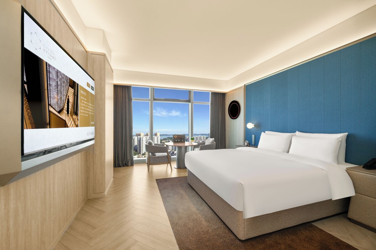 Une chambre d’hôtel moderne avec un grand lit, un téléviseur fixé au mur et une vue sur le paysage urbain.