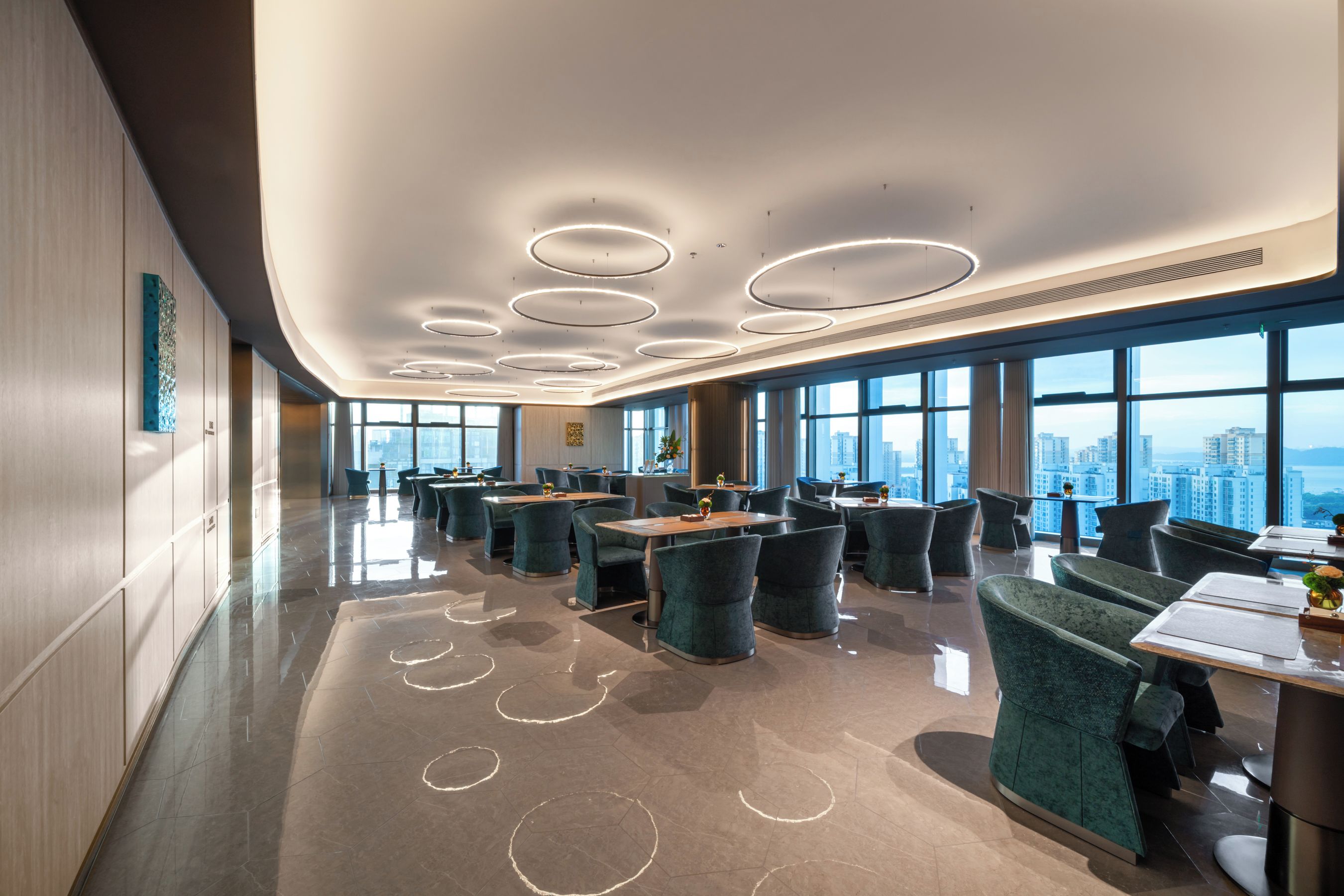 Un'area pranzo moderna con soffitto alto, lampade circolari e ampie finestre che offrono vista sulla città.