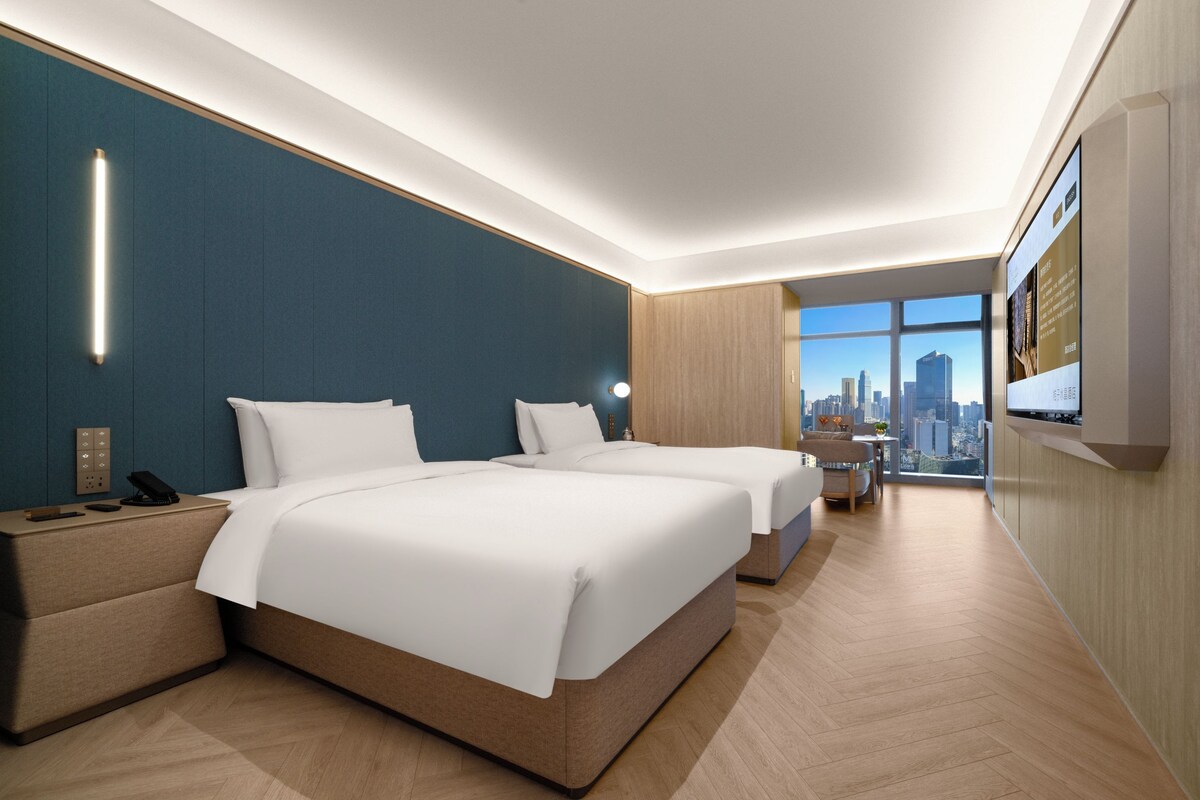 A hotel room with two beds, a desk, a chair, a TV, and a city view.