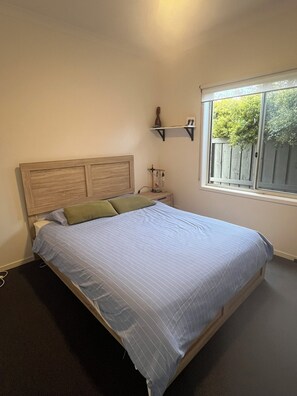 A bedroom with a wooden headboard, a bed with striped bedding, a bedside table with a lamp, and a window with a view of greenery. - Your Comfortable, Spacious Phillip Island Getaway (Cowes)