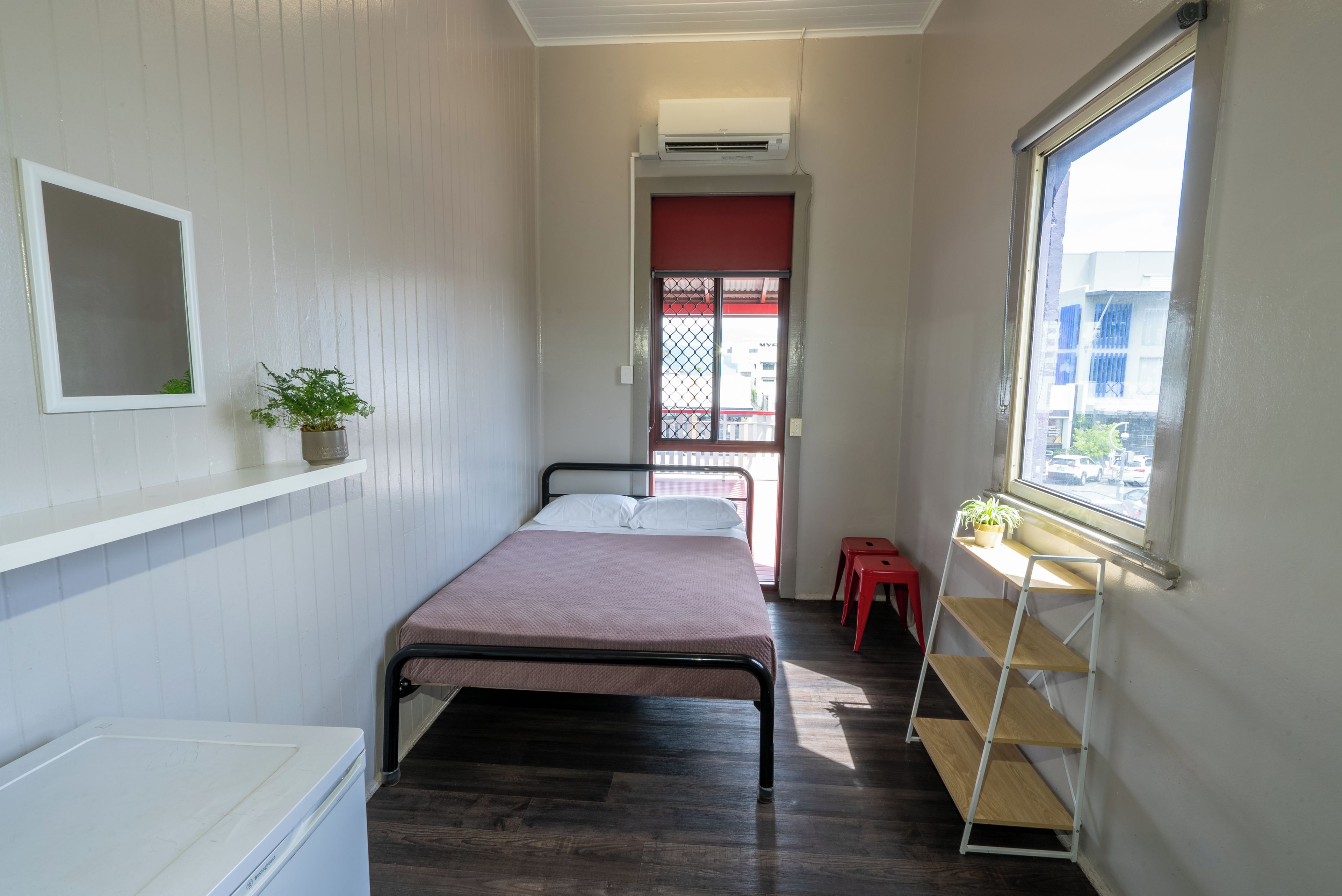A small, narrow room with a bed, a shelf with a plant, a window with a view of buildings, and a red door.