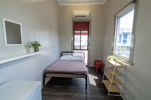 A small, narrow room with a bed, a shelf with a plant, a window with a view of buildings, and a red door. - The Jack Backpackers (Cairns)