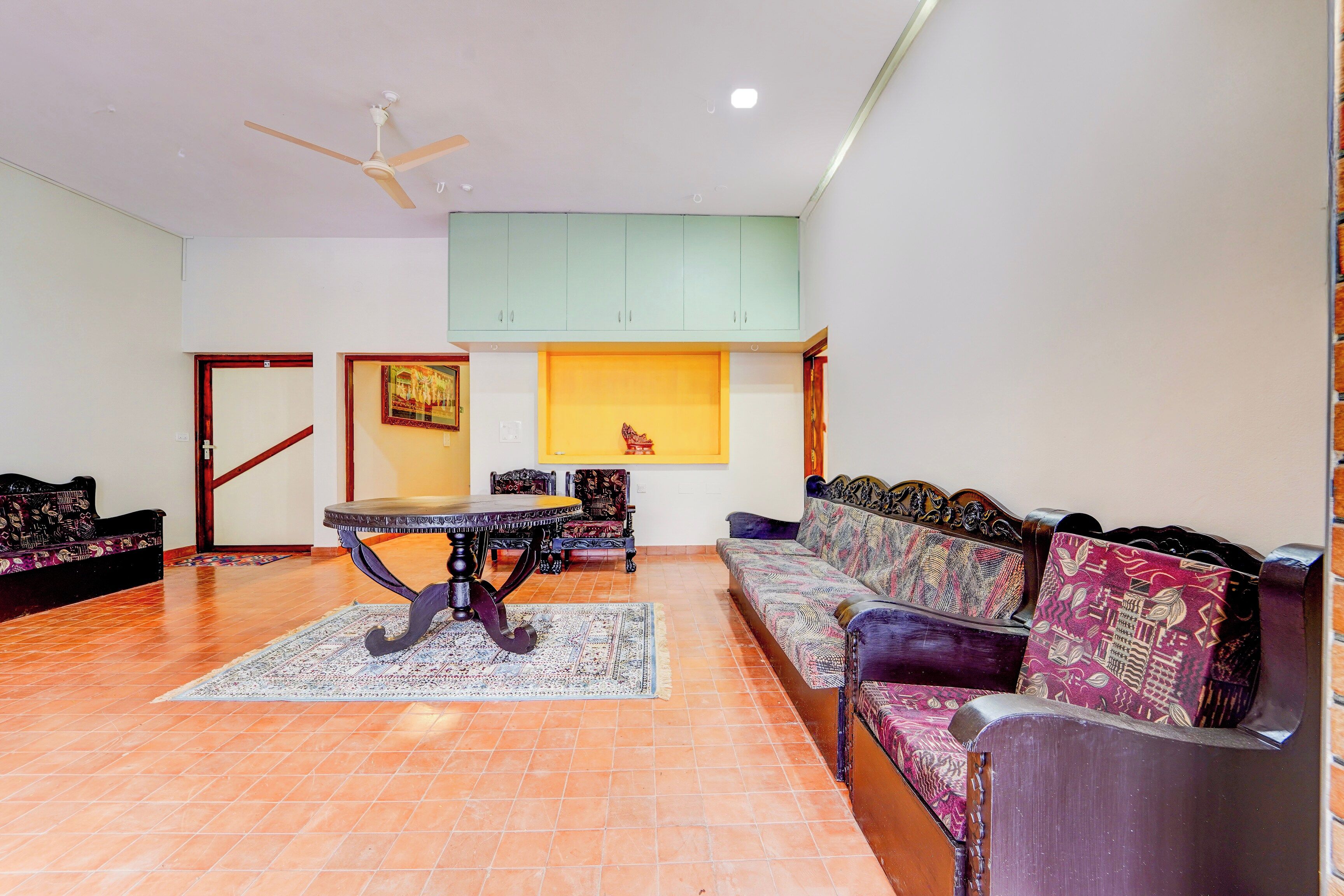 A spacious living room with a high ceiling, a ceiling fan, a dining table with chairs, a sofa set, and a patterned rug.