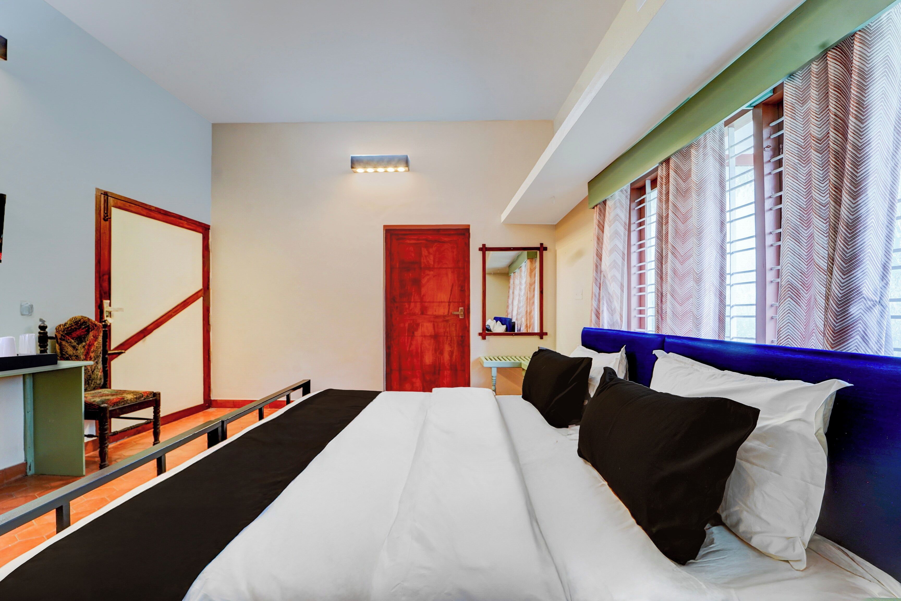 A hotel room with a large bed, a wooden chair, a desk with a mirror, and a window with patterned curtains.