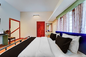 Ett hotellrum med en stor säng, en trästol, ett skrivbord med en spegel och ett fönster med mönstrade gardiner.