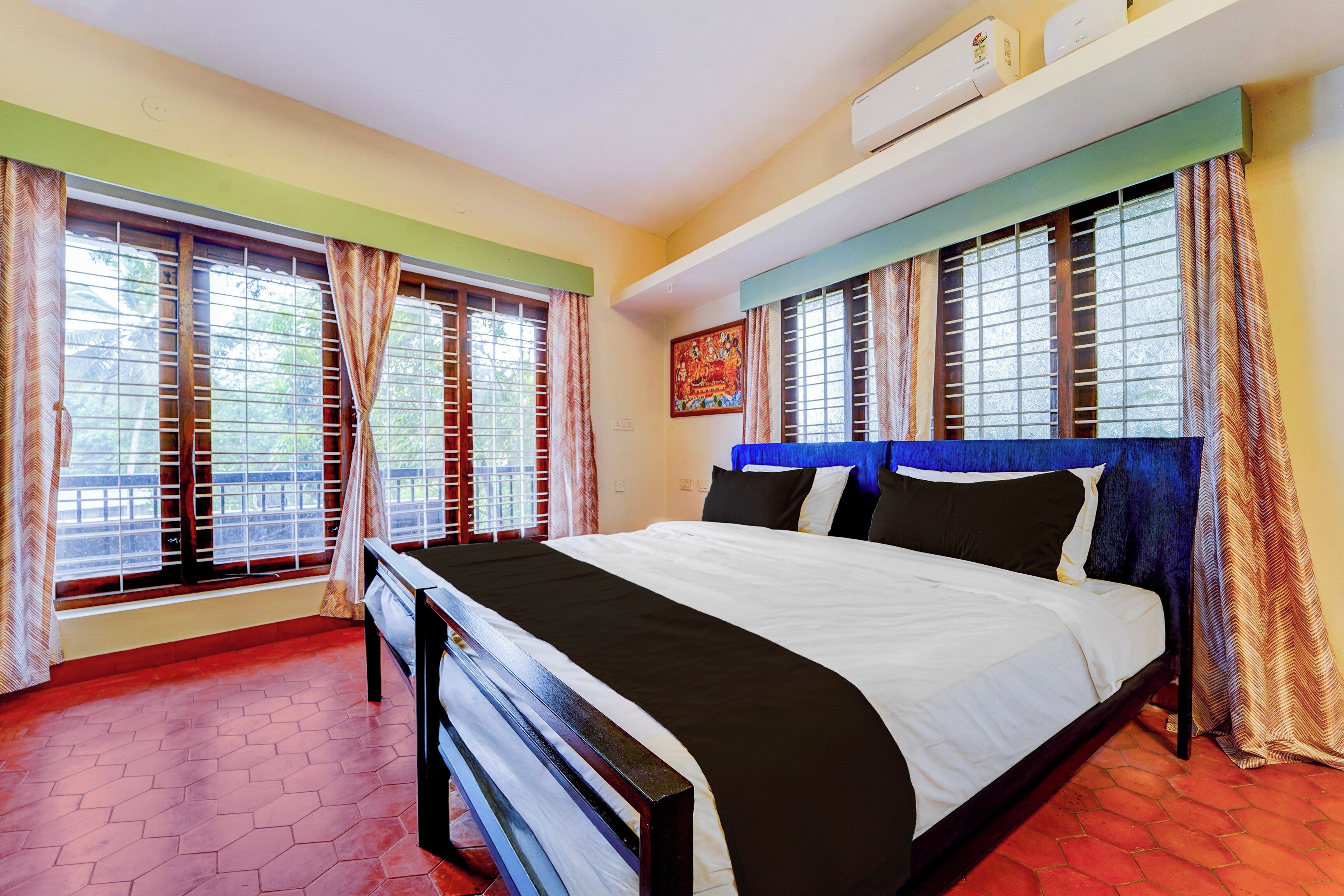 A hotel room with a bed, two windows with curtains, an air conditioning unit, and a painting on the wall.
